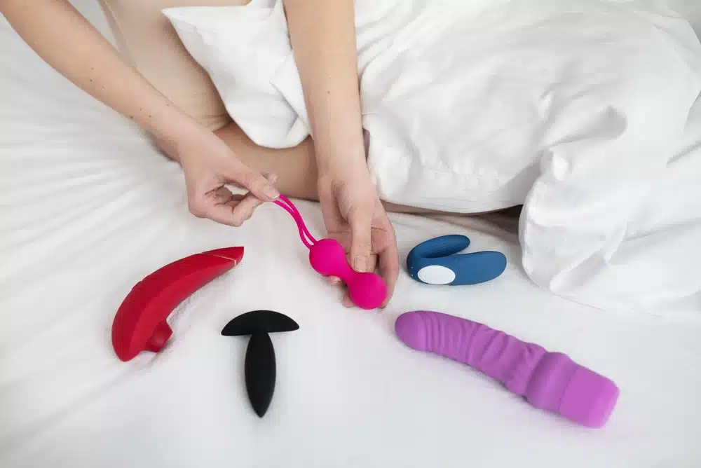 egy ágyon szexjátékok láthatóak és egy nő keze, amint egy piros kegel golyót tart